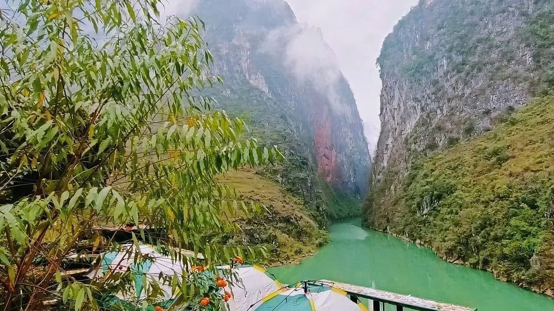 Tents on a riverside deck above the emerald Nho Que River, with misty limestone cliffs on both sides of the canyon