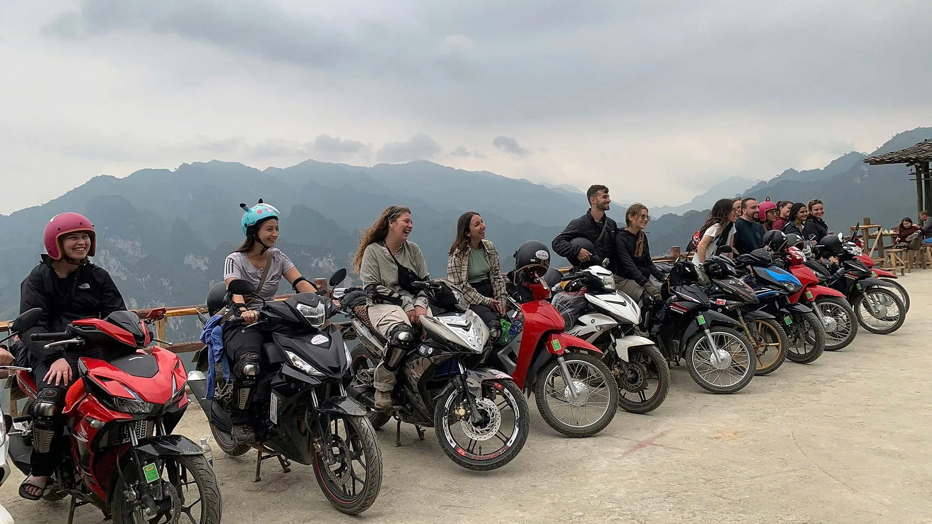 Riders from a Freedom group sitting on their parked motorbikes at a mountain viewpoint on a cloudy day in Ha Giang